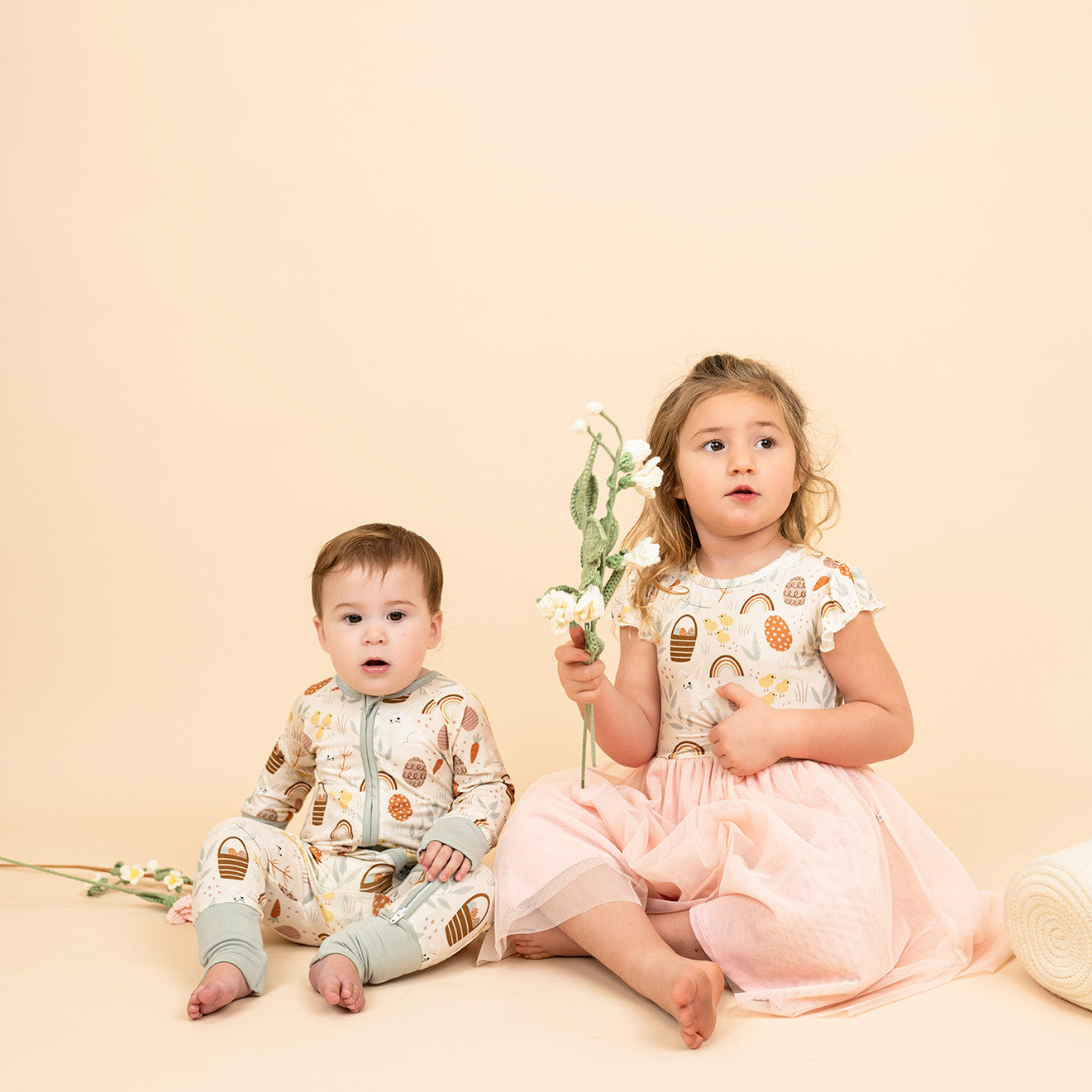 Two children sitting down in our Hop to It print. One child is in our convertible pajamas and the other child is in our tulle dress holding flowers