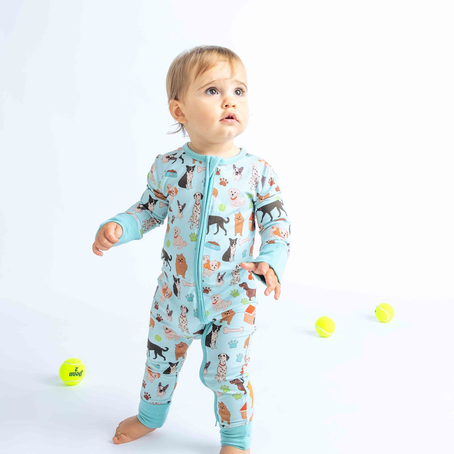 Baby standing modeling bamboo zippy pajama. Print is blue featuring different types of dogs, a dog house, paw prints, bones, and more! 