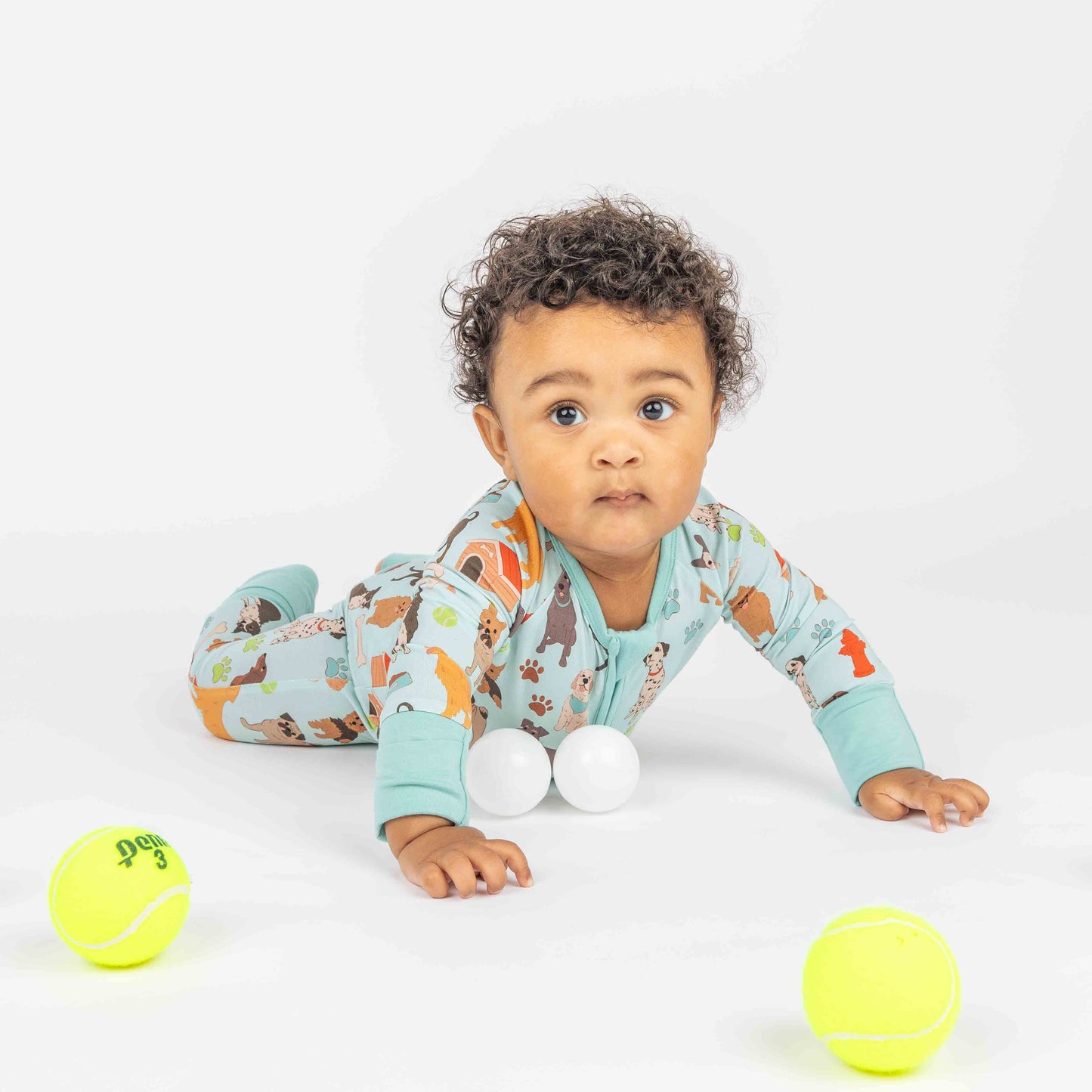 Baby is laying on white background with balls around modeling zippy pajama. Print is blue featuring different types of dogs, a dog house, paw prints, bones, and more! 