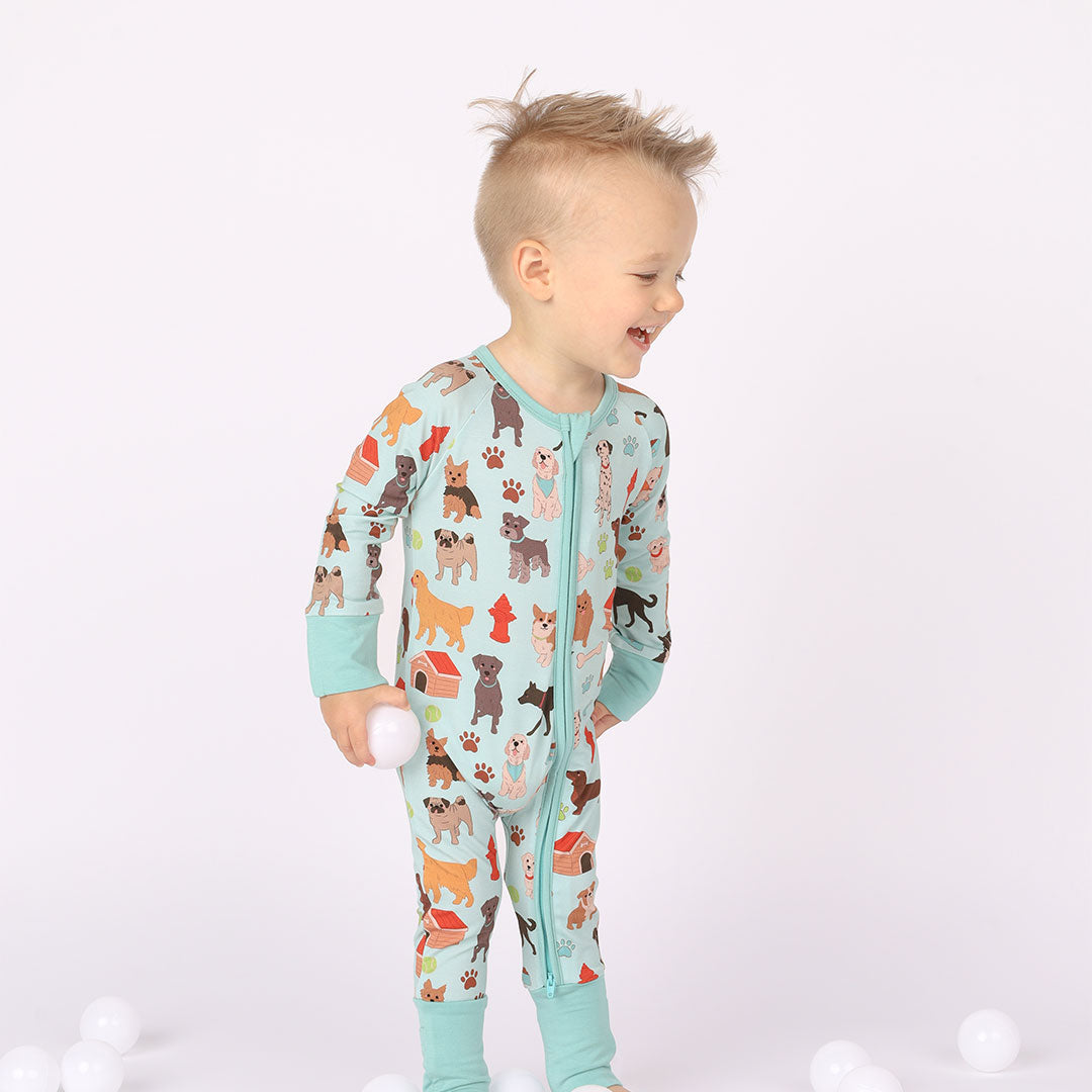 Boy is smiling modeling baby zippy pajama. Print is blue featuring different types of dogs, a dog house, paw prints, bones, and more! 