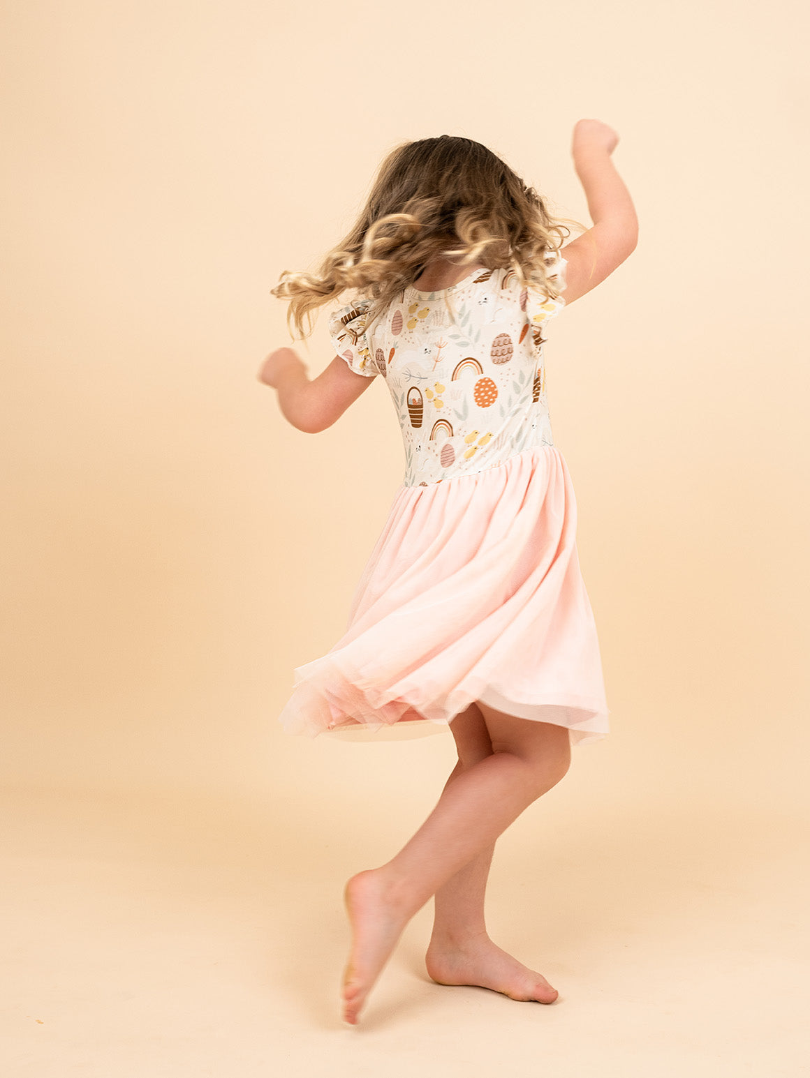 Girl twirling around in her Hop to It Tulle Dress by Emerson and Friends. The print hop to it features bunnies, easter eggs, chicks and more!