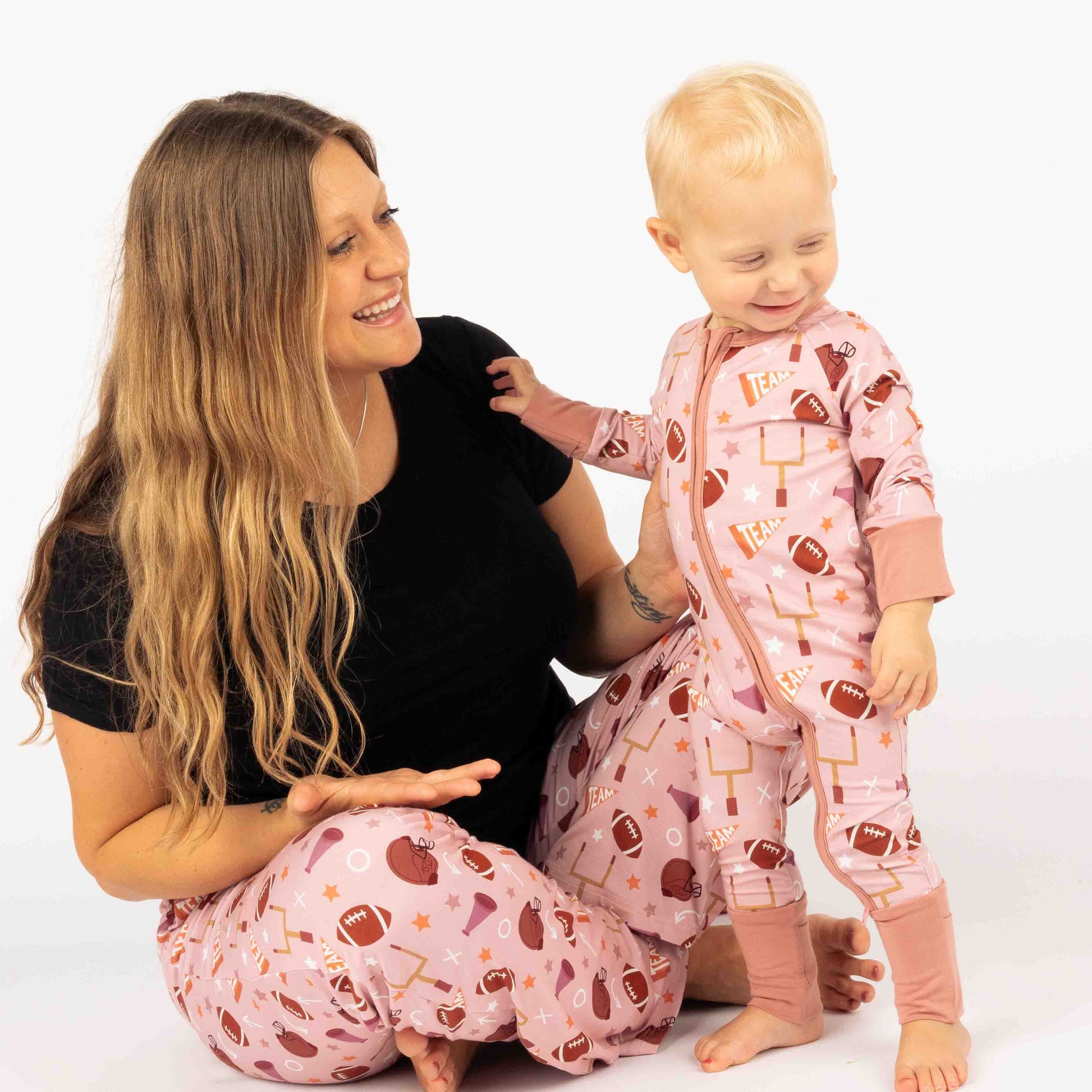 Mom smiling while holding her baby girl sitting on white backdrop. Both wearing our pink football print called "Game Day" Featuring footballs, goal posts, stars and more! 