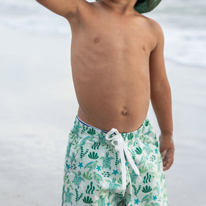 Boy int the beach wearing our Save the Sea Turtle Swim trunks. The print is featuring a sea-green turtle and ocean-inspired print. Made from quick-dry, lightweight fabric with an adjustable drawstring waist for a comfy fit. Perfect for beach days and poolside fun!