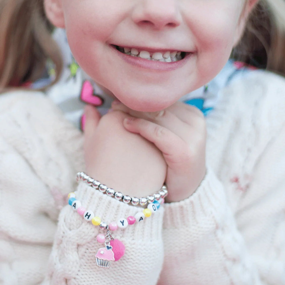 Young girl outside wearing a colorful beaded bracelet with letter beads spelling Happy Birthday, a pink pom-pom and a pink frosted cupcake charm from Great Pretenders 