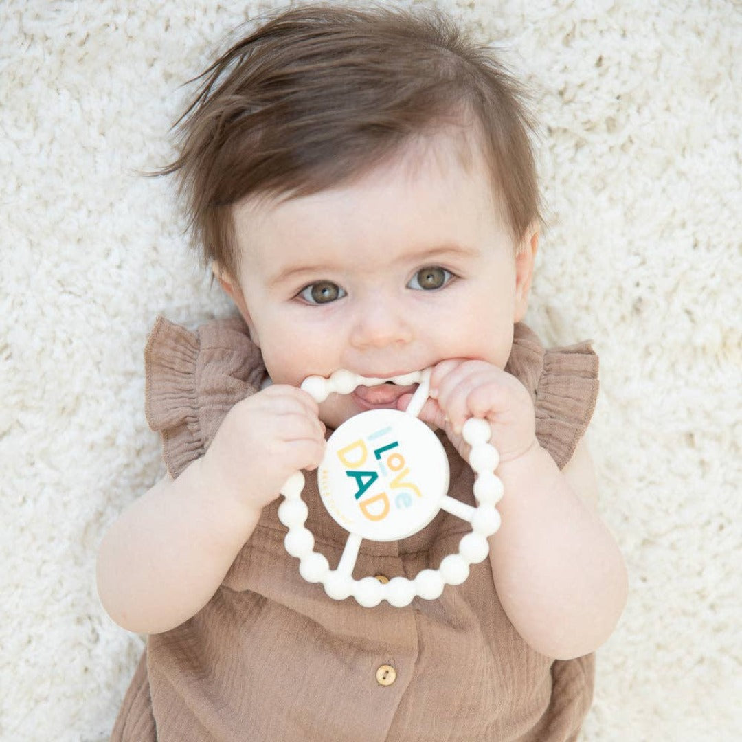 little baby chewing on a white round teething toy that says I love dad in multiple colors