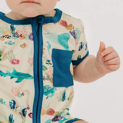 Baby girl close up wearing our Emerson and Friends Coral Reef Bamboo Baby Shortie Romper, a soft and breathable summer outfit featuring an ocean-inspired print with sharks, sea turtles, and coral, designed for all-day comfort with an easy zip closure.