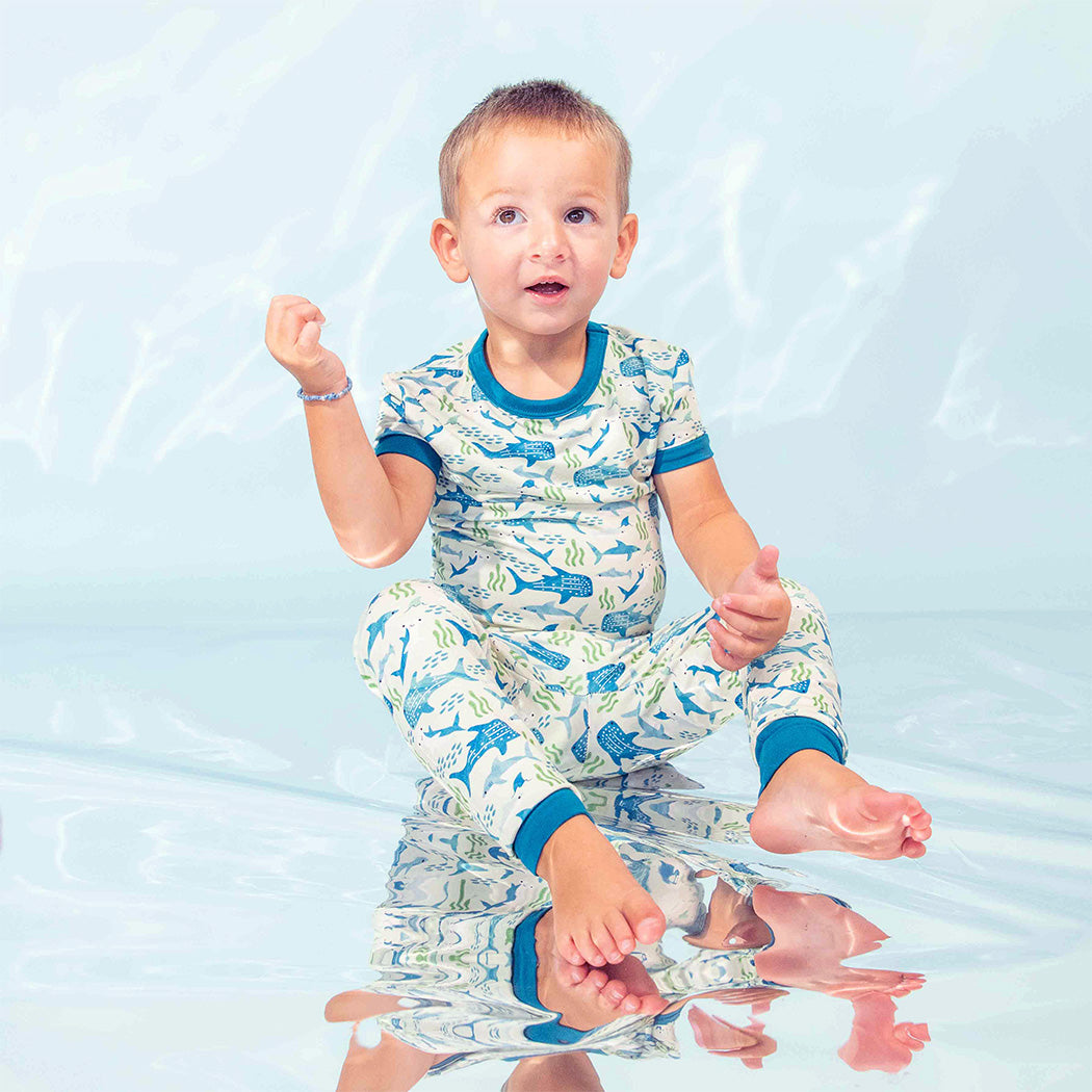 Boy wearing our Shark Friends Two-piece Bamboo Pajamas sitting on the metallic-under-water like backdrop. 