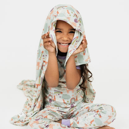Little girl wearing her Magical Unicorn pajama set, with our Magical Unicorn blanket over her head. The print includes unicorns, rainbows, clouds, planets, stars, and hearts. The colors are purple, pink, and blu, with hints of green. 