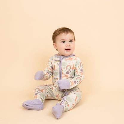 Happy baby sitting while wearing our Magical Unicorn print with foldable hand and foot covers. The print includes unicorns, rainbows, clouds, planets, stars, and hearts. The colors are purple, pink, and blu, with hints of green. 