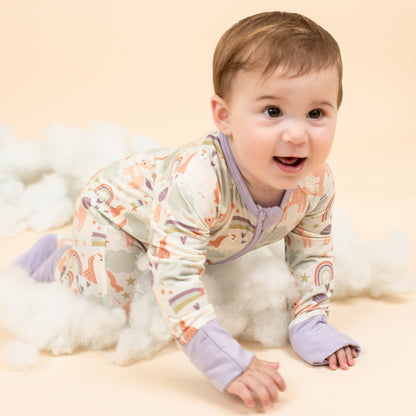 Little baby girl crawling on her hands and knees in our Magical Unicorn baby bamboo convertible pajama. The print includes unicorns, rainbows, clouds, planets, stars, and hearts. The colors are purple, pink, and blu, with hints of green. 