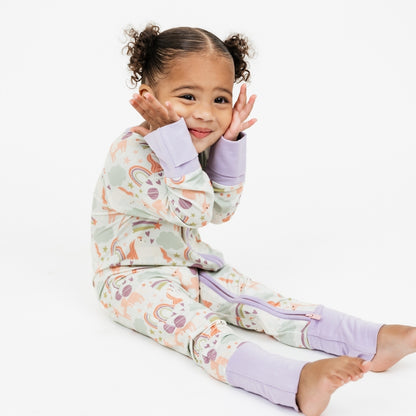 Little girl sitting down in our Magical Unicorn bamboo convertible pajama with her hands on her face smiling. The print includes unicorns, rainbows, clouds, planets, stars, and hearts. The colors are purple, pink, and blu, with hints of green. 