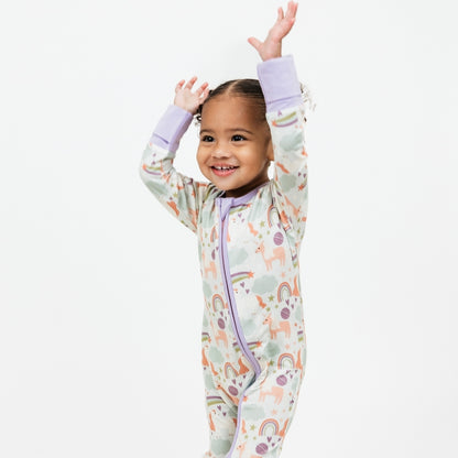 Happy toddler throwing her hands up in excitement while wearing our Magical Unicorn convertible pajama. The print includes unicorns, rainbows, clouds, planets, stars, and hearts. The colors are purple, pink, and blu, with hints of green. 