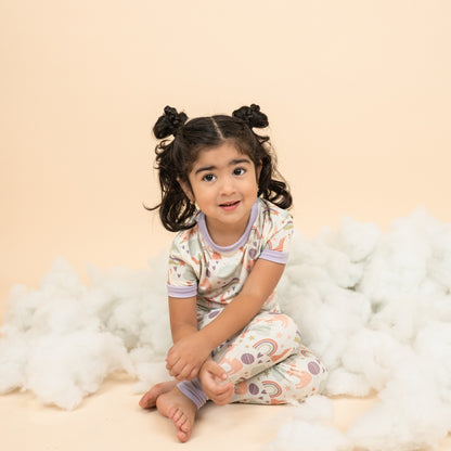Little girl wearing our Magical Unicorn bamboo toddler pajama set atop fake clouds. The print includes unicorns, rainbows, clouds, planets, stars, and hearts. The colors are purple, pink, and blu, with hints of green. 