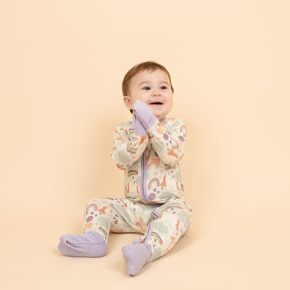 Excited little girl in our Magical Unicorn bamboo convertible pajama clapping hands together. Her feet and hands have the foldable covers on them from the pajama. The print includes unicorns, rainbows, clouds, planets, stars, and hearts. The colors are purple, pink, and blu, with hints of green. 