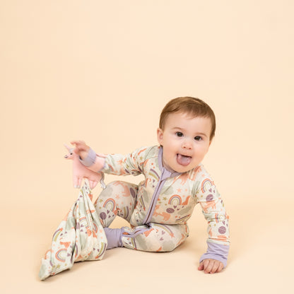 Little girl wearing our Magical Unicorn convertible holding the Magical Unicorn Lovey. The print includes unicorns, rainbows, clouds, planets, stars, and hearts. The colors are purple, pink, and blu, with hints of green. 