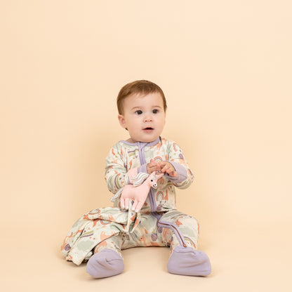 Little girl wearing our Magical Unicorn convertible holding the Magical Unicorn Lovey. The print includes unicorns, rainbows, clouds, planets, stars, and hearts. The colors are purple, pink, and blu, with hints of green. 