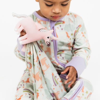 Little girl wearing our Magical Unicorn convertible holding the Magical Unicorn Lovey. The print includes unicorns, rainbows, clouds, planets, stars, and hearts. The colors are purple, pink, and blu, with hints of green. 