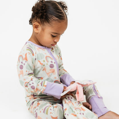 Little girl wearing our Magical Unicorn convertible holding the Magical Unicorn Lovey. The print includes unicorns, rainbows, clouds, planets, stars, and hearts. The colors are purple, pink, and blue, with hints of green. 