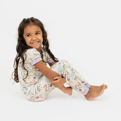 Little girl sitting down with her arms under her legs, wearing our Magical Unicorn bamboo pajama set. The print includes unicorns, rainbows, clouds, planets, stars, and hearts. The colors are purple, pink, and blu, with hints of green. 