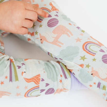 Zoomed in detailed picture of the girl pointing to her leg as she wears our Magical Unicorn bamboo pajama set. The print includes unicorns, rainbows, clouds, planets, stars, and hearts. The colors are purple, pink, and blu, with hints of green. 