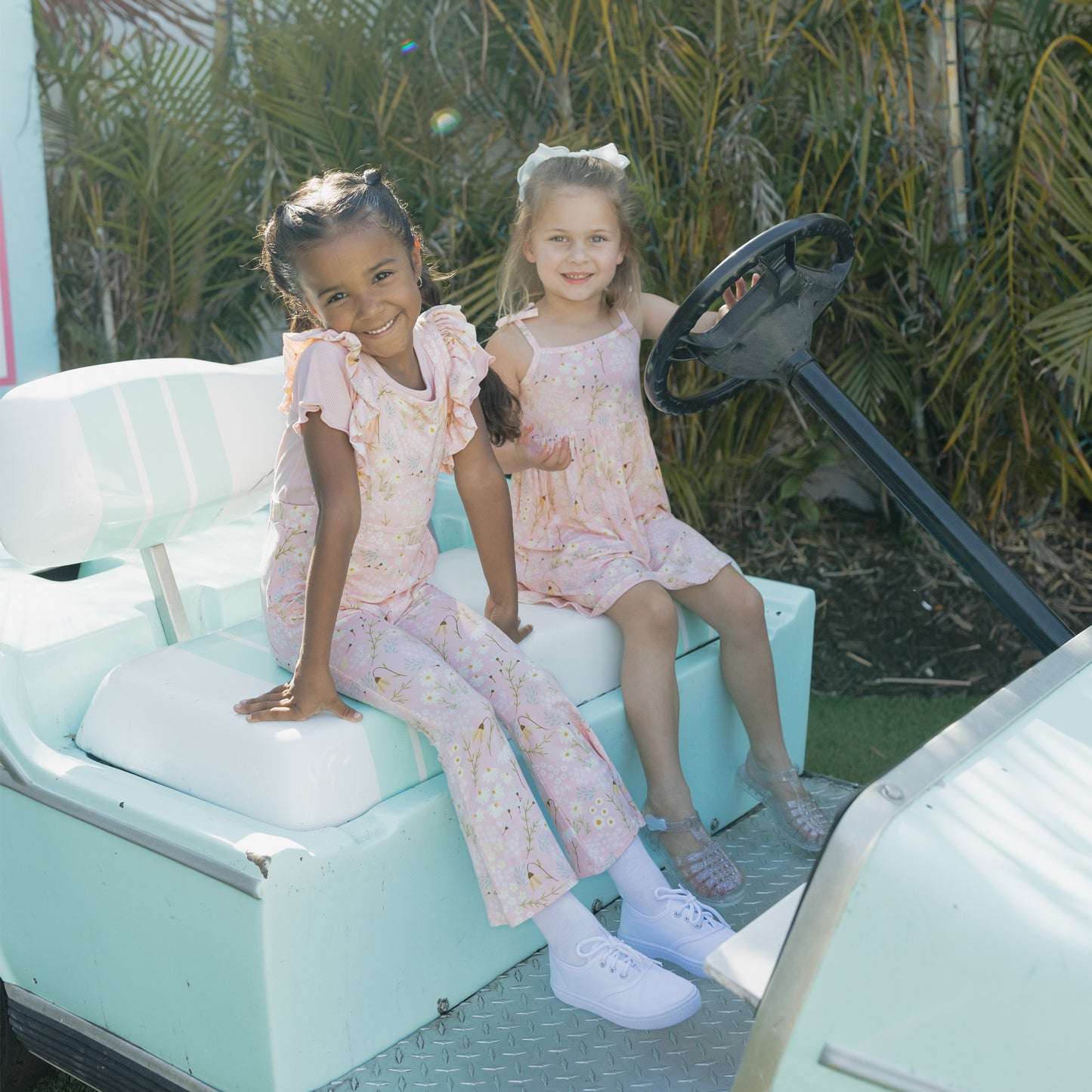 Two young girls in matching outfits sitting in a light blue vehicle with a green outdoor background.