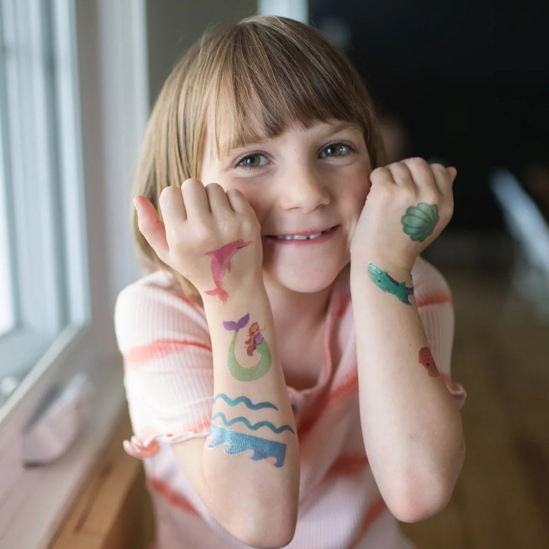 Child with colorful mermaid and aquatic animals temporary tattoos on her arms sitting by a window from Great Pretenders 