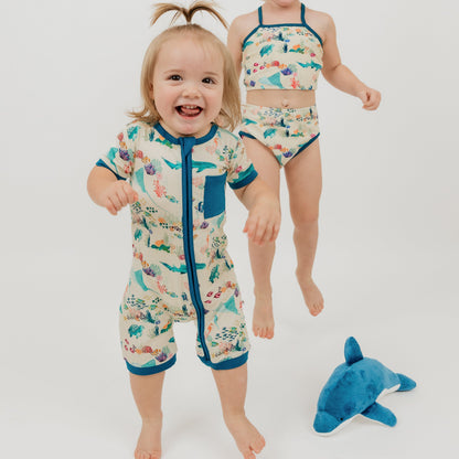 Two toddler wearing our Emerson and Friends Coral Reef Bamboo Baby Shortie Romper and our 2 piece swimwear, a soft and breathable summer outfit featuring an ocean-inspired print with sharks, sea turtles, and coral, designed for all-day comfort with an easy zip closure.