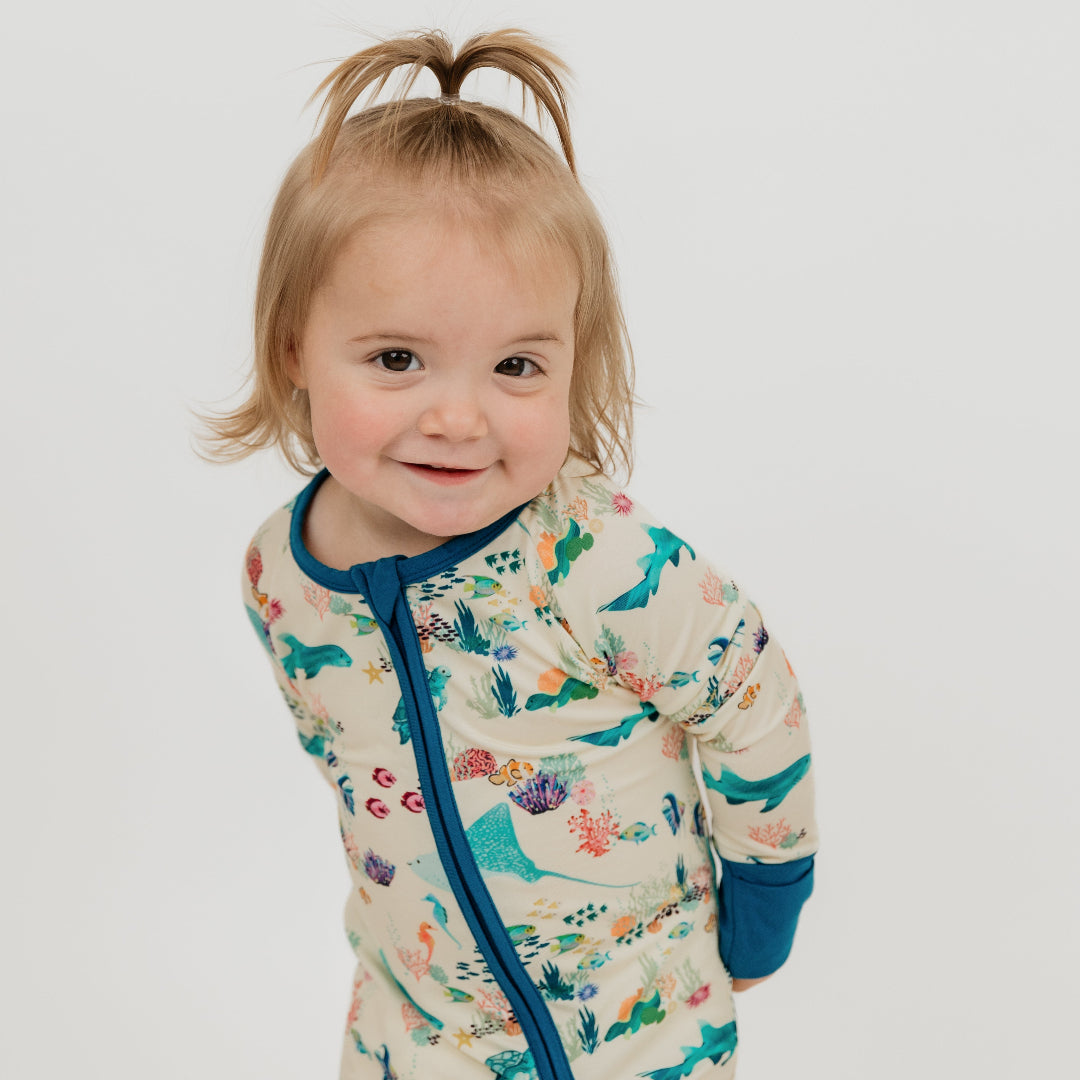 Baby girl dressed in a coral reef bamboo convertible with vibrant ocean design, soft blue trim, and a two-way zipper for quick changes.