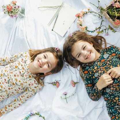Two young girls lying on a white blanket, smiling at the camera. The girl on the left wears a cream floral long-sleeve top, and the girl on the right wears a dark teal floral long-sleeve top.