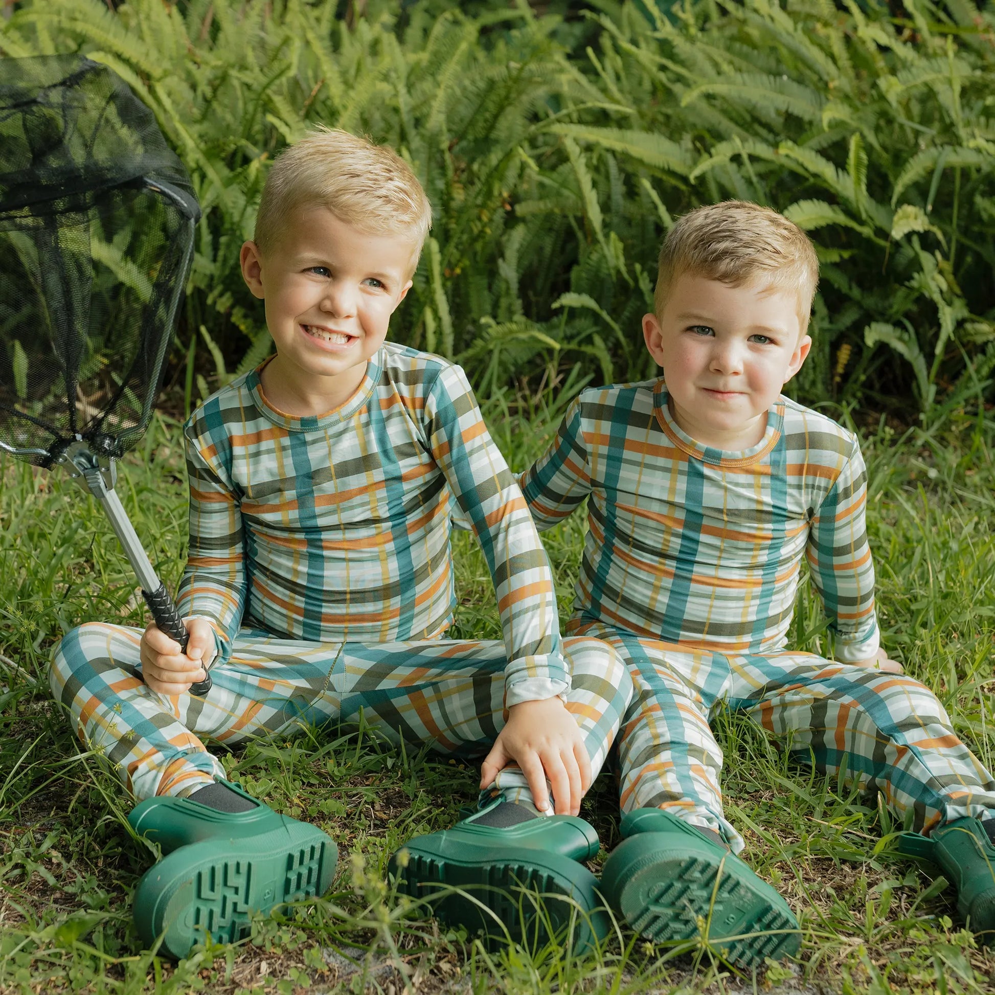 two boys wearing a fall color plaid pajama set on greenery background. 