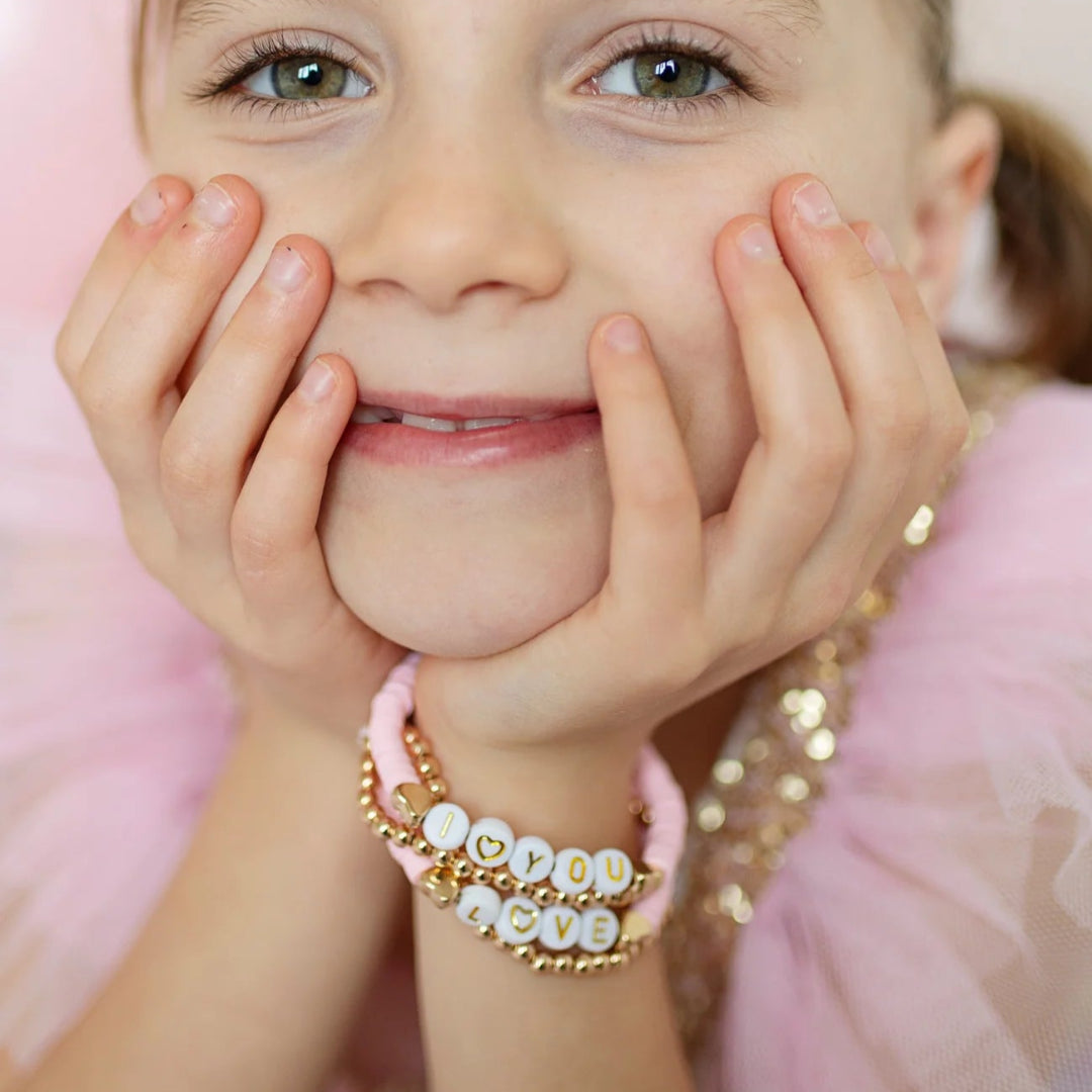 Young girl with hands on cheeks wearing four stacking bracelets that are pink and gold with white letter beads spelling out "I love you" and "Love" from Great Pretenders