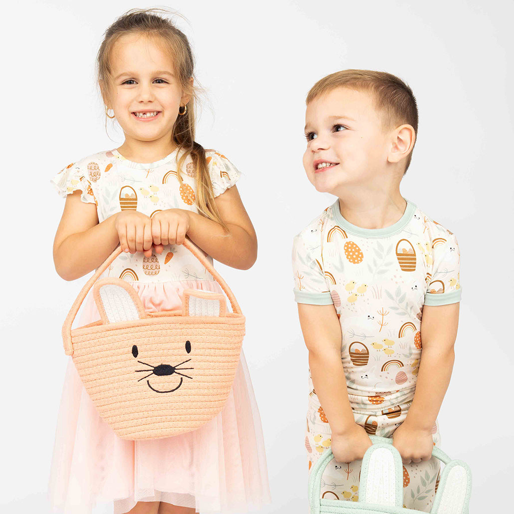 Two toddlers smiling while showcasing our bamboo products. The girl is wearing our Easter dress and holding a pink bunny basket, while the boy is dressed in a two-piece bamboo pajama set.
