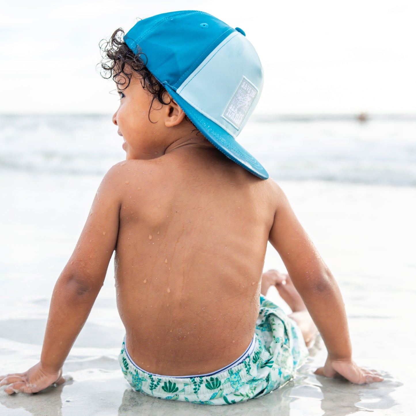 A young boy sitting on the beach wearing pieces from our Save the Sea Turtle collection, including the hat and matching swimwear — a perfect blend of style and ocean advocacy.