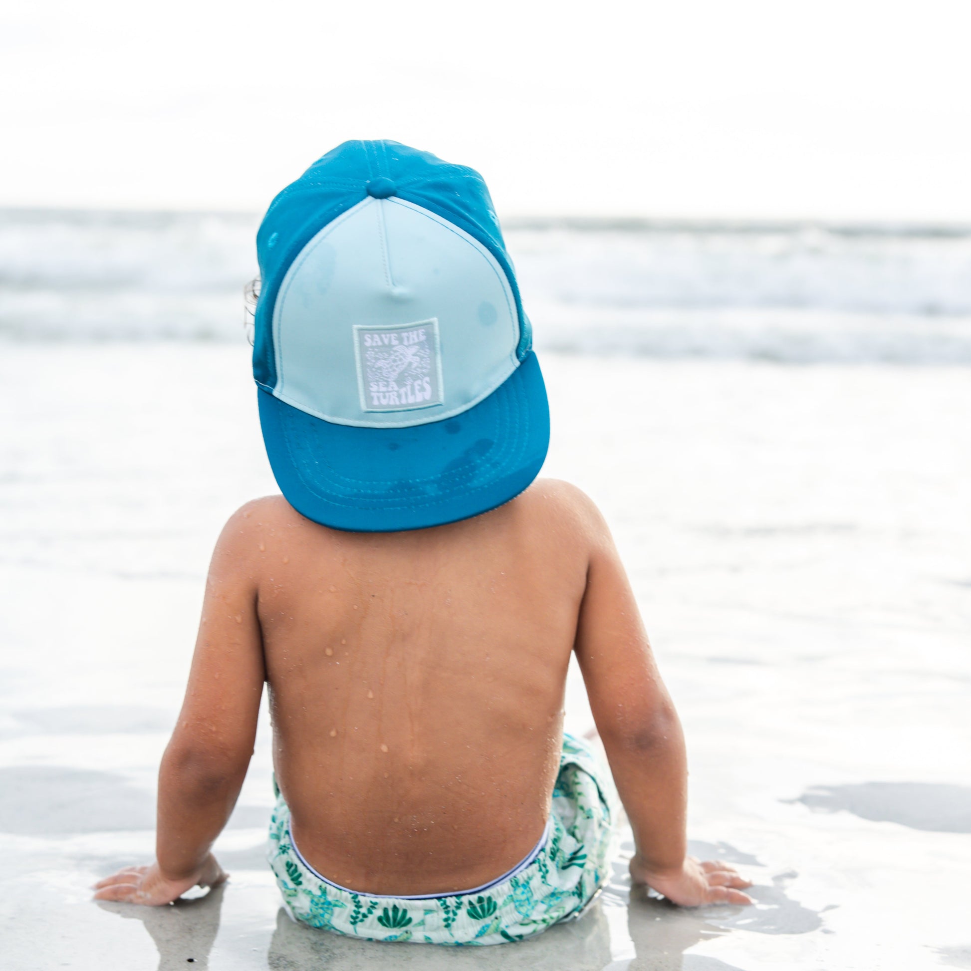 A young boy sitting on the beach wearing pieces from our Save the Sea Turtle collection, including the hat and matching swimwear — a perfect blend of style and ocean advocacy.