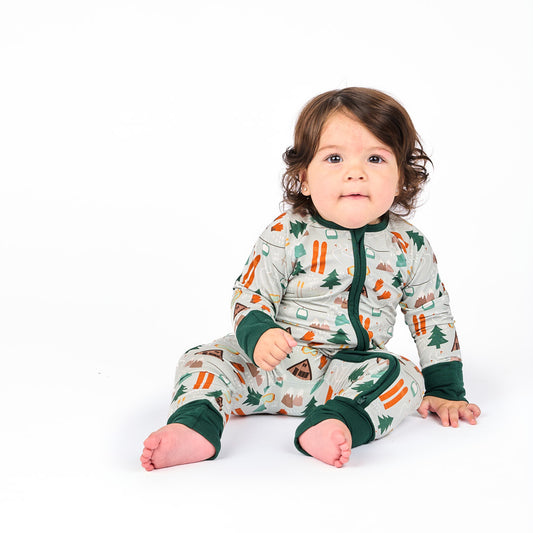 Baby sitting in white background wearing convertible bamboo pajama ski day featuring trees, cabins, and other ski activities.