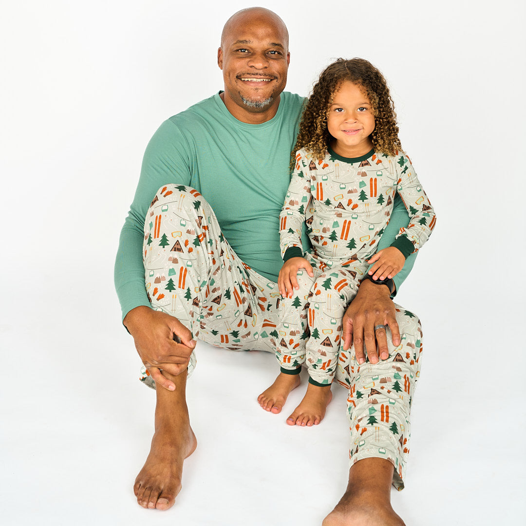 Father and daughter modeling in white background wearing bamboo pajama ski day featuring trees, cabins, and other ski activities.