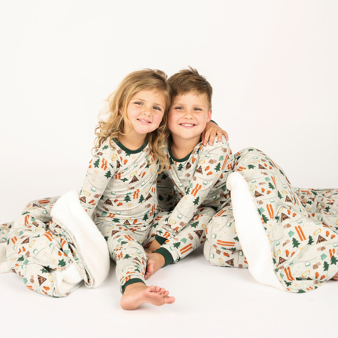 Siblings modeling in white background wearing bamboo pajama ski day featuring trees, cabins, and other ski activities and wearing a blanket