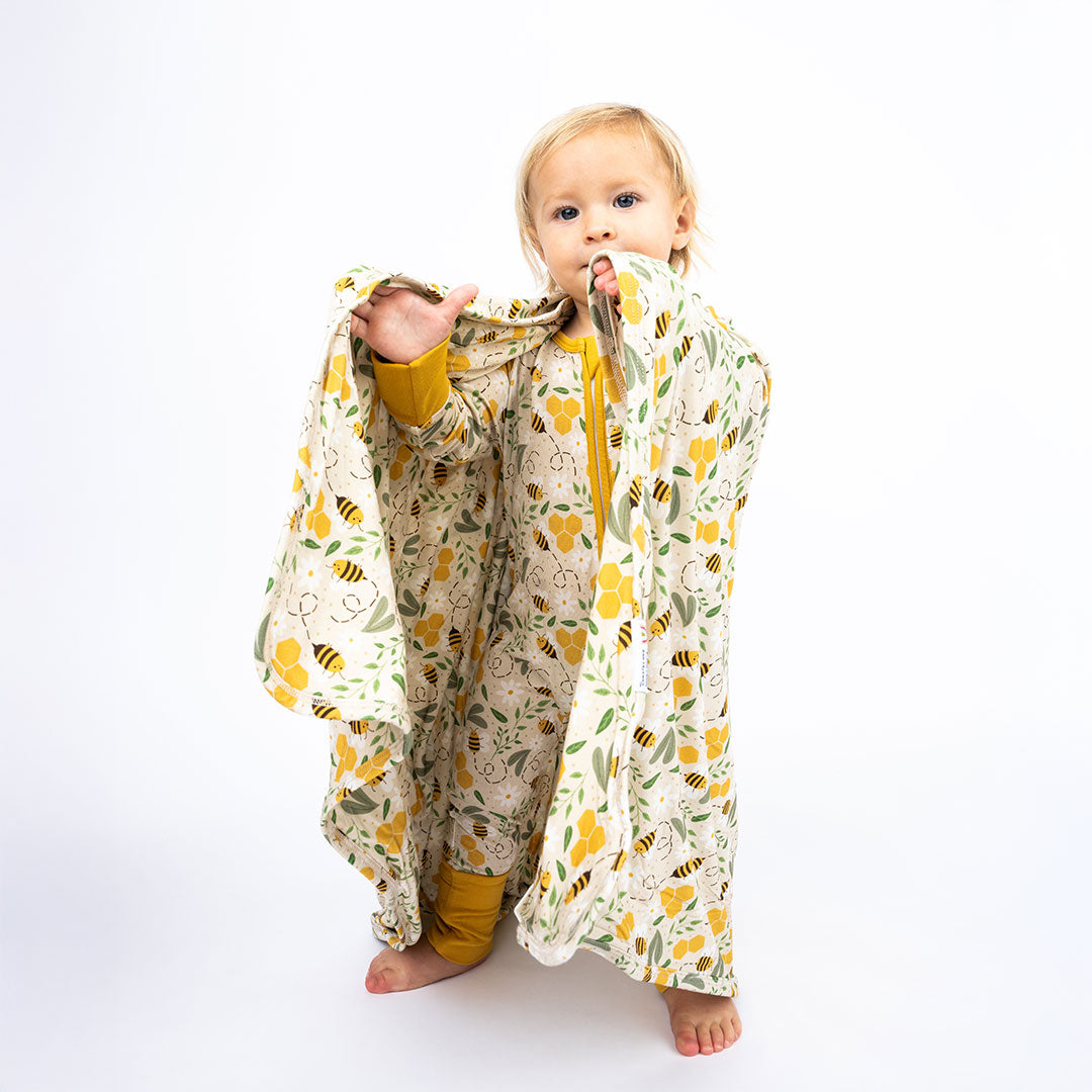 Baby boy holding our luxury bamboo Happy Honey Bee Blanket on white background. Print has bumble bees zooming around with green leaves, white flowers and honeycombs.
