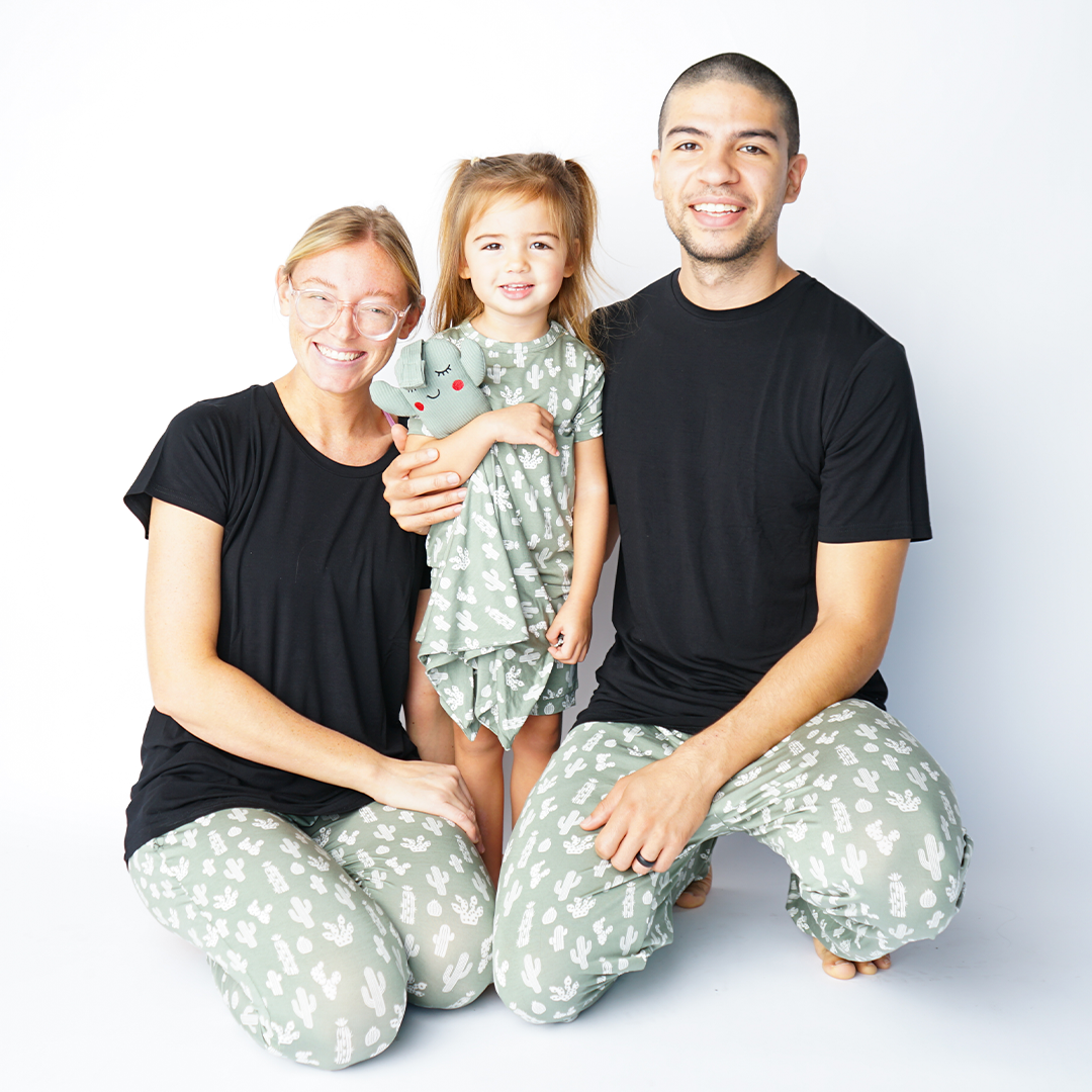 family of 3 smiles in their "stay sharp apparel. Mom and dad sit on the floor and hold on to their baby who stands. mom  is in the jogger pants, dad wears the relaxed pants, and the baby wears the convertible. the "stay sharp" print is a variety of different white cacti on a greyish/green background. 