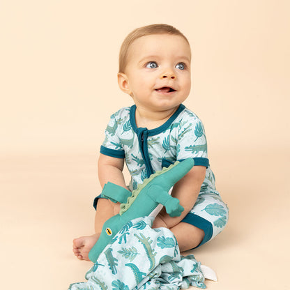 smiling baby in his "later gator" short. he is playing with his "later gator" lovey. the "later gator" print has a mix of light and dark green alligators, leaves, and white dots scattered on a teal colored background. 