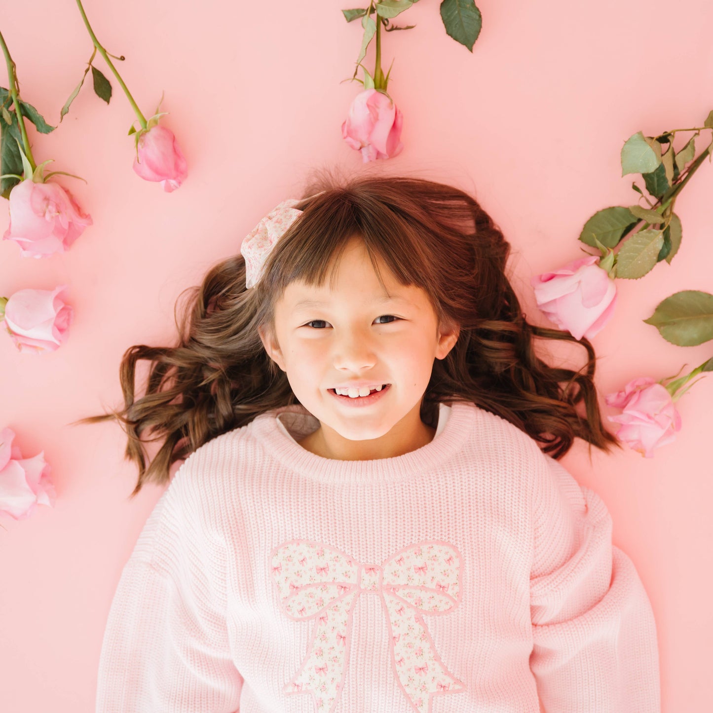Young girl in a pink sweater with a bow, lying on a pink background with flowers.