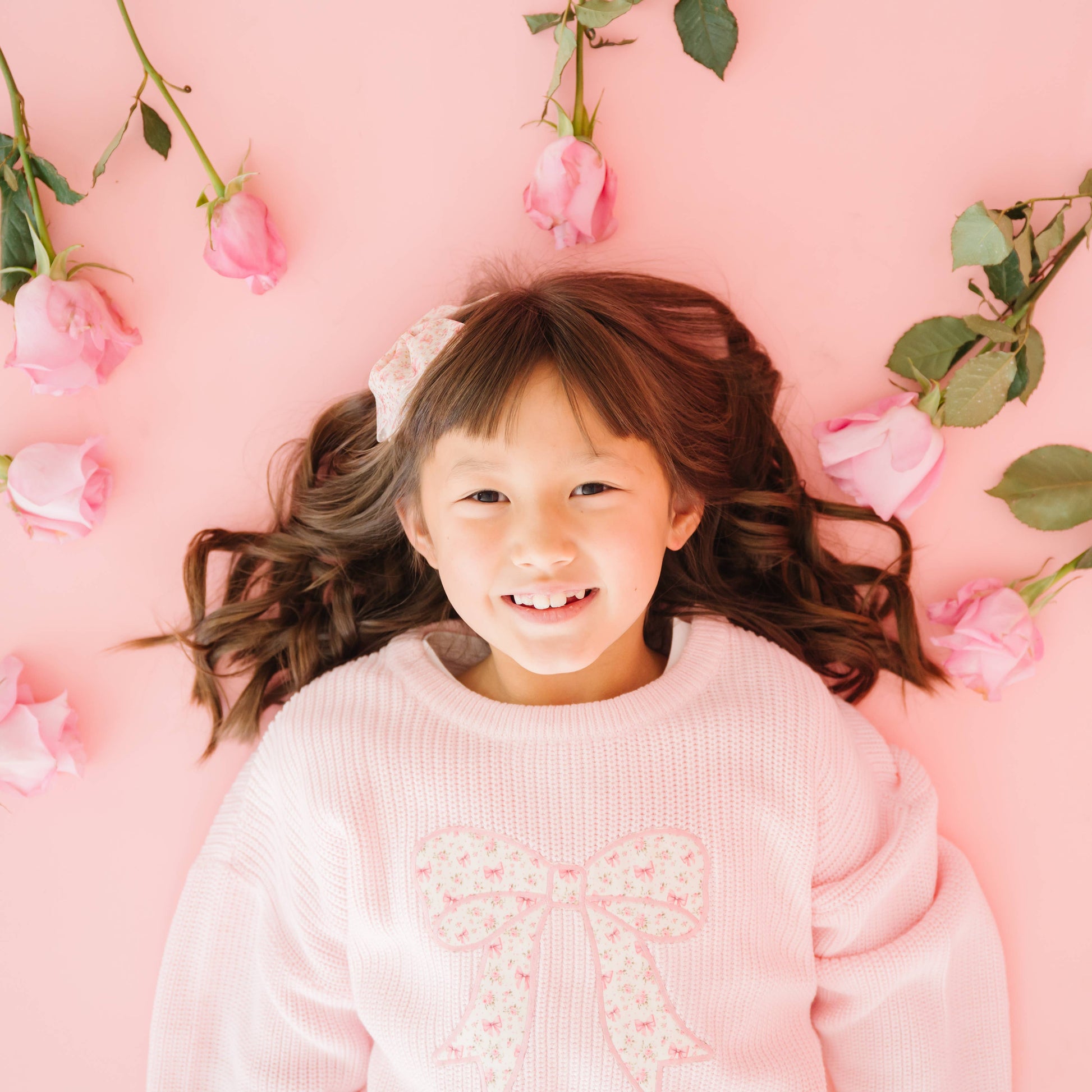 Young girl in a pink sweater with a bow, lying on a pink background with flowers.