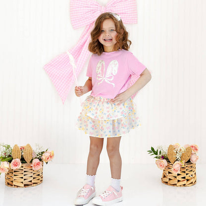 Young girl in a pink outfit with floral skirt and shoes, holding a pink umbrella on a white background.