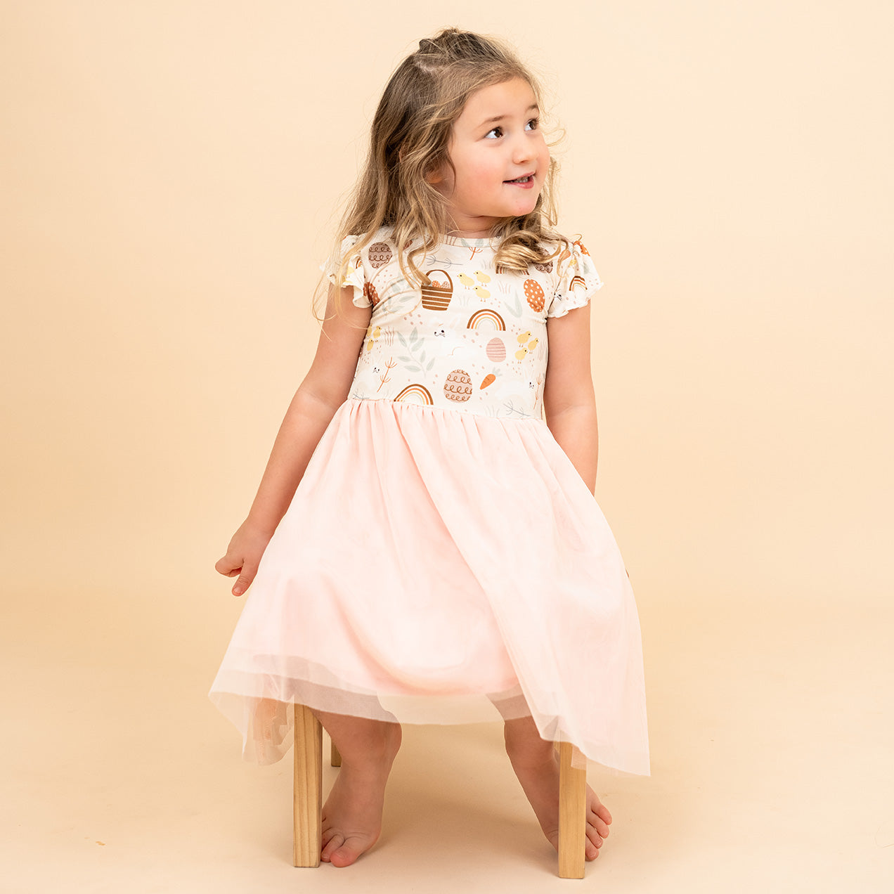 Little girl sitting on a chair with her tulle bamboo dress. The print he has on is our Hop to it dress that features bunnies, chicks, easter eggs and more!