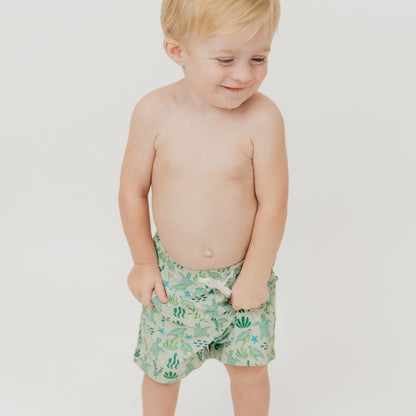 Smiling boy wearing our Save the Sea Turtle Swim trunks. The print is featuring a sea-green turtle and ocean-inspired print. Made from quick-dry, lightweight fabric with an adjustable drawstring waist for a comfy fit. Perfect for beach days and poolside fun!
