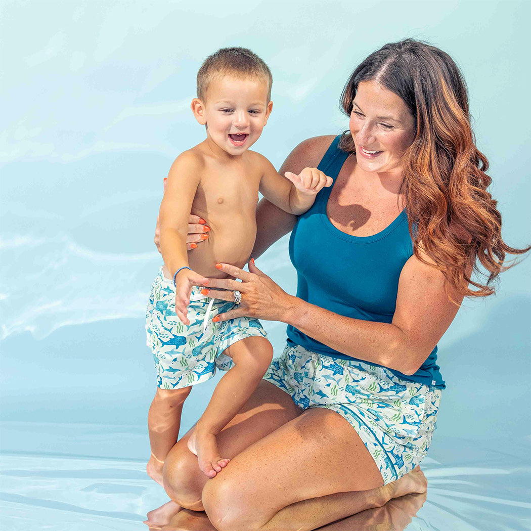 Mom wearing our Shark Friends Bamboo Pajama Shorts and Women's Ocean Blue Tank Top, holding her son while he's wearing the Shark Friends Swim Trunks. 