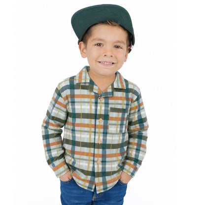 A smiling young boy wearing a green cap, a long-sleeve plaid button-up shirt in green, beige, and orange tones, and blue jeans, photographed against a plain white background.