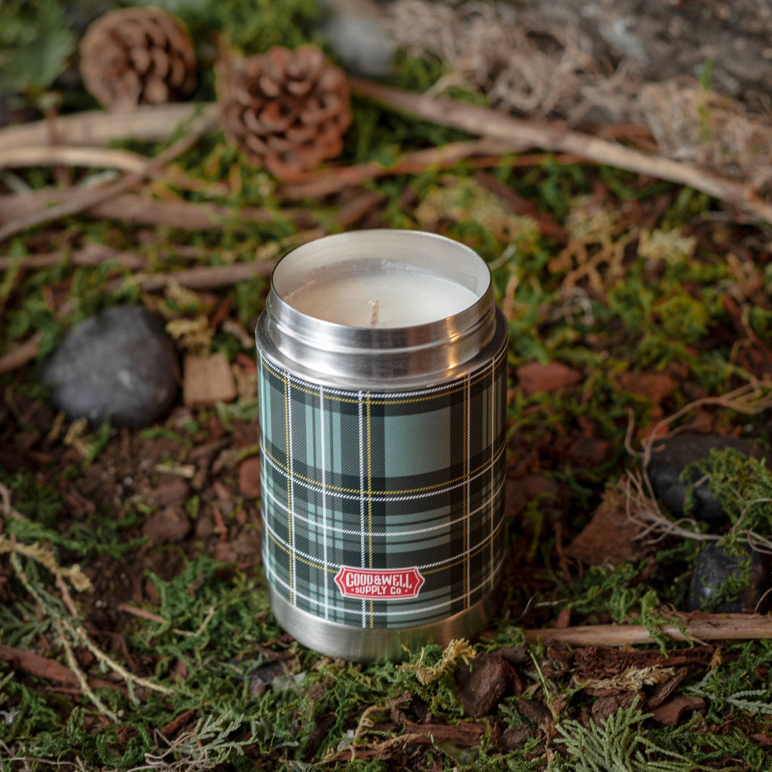 Candle in a plaid container on a natural background with pine cones and leaves.