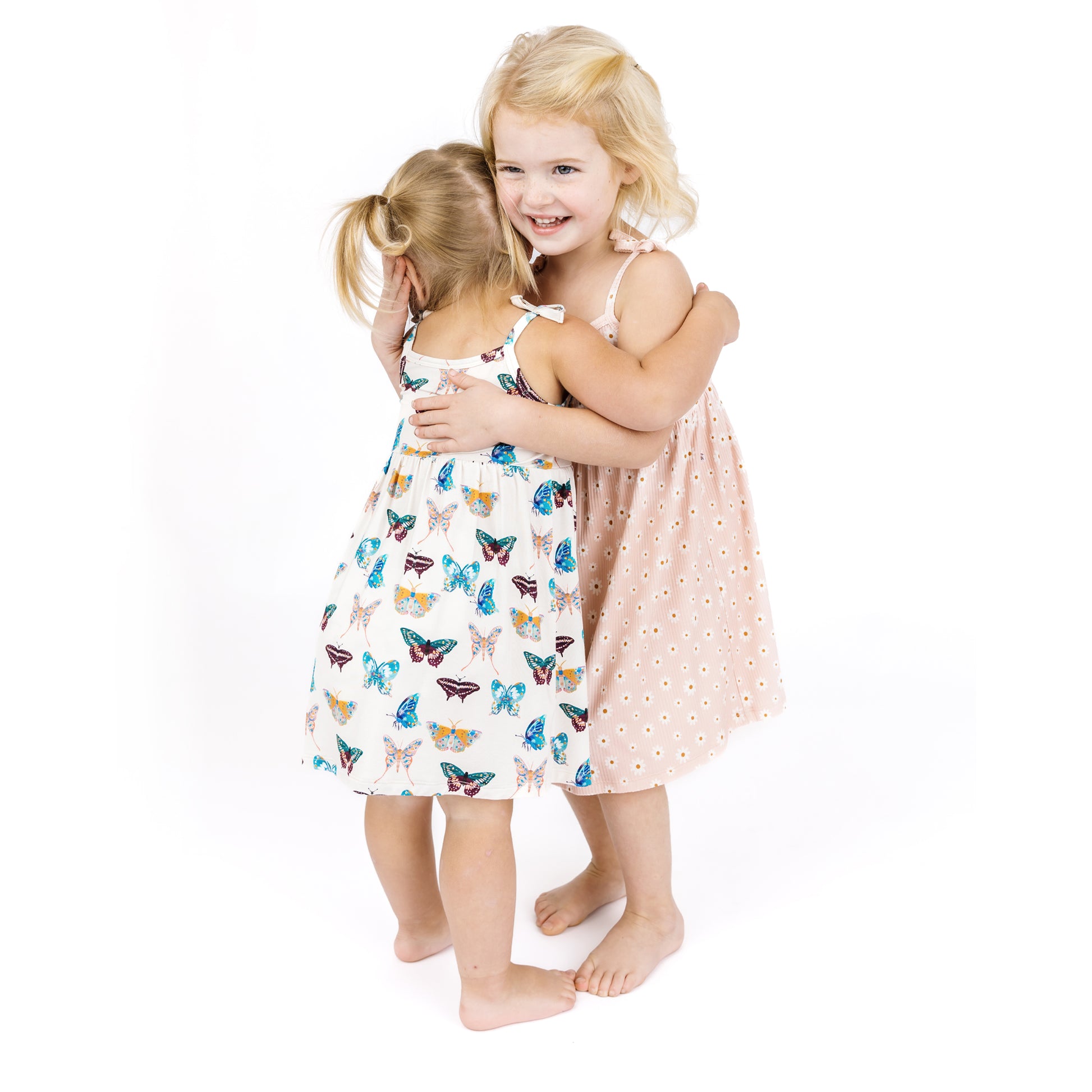 Two young girls hugging each other wearing dresses with butterfly and pink daisy patterns on a white background.