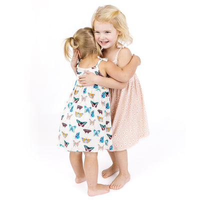 Two young girls hugging each other wearing dresses with butterfly and pink daisy patterns on a white background.